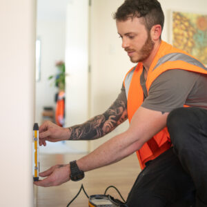 This image features a tradesperson wearing a bright orange safety vest and working indoors. They are using a measuring tool on a wall, with a focus on precision and care. The worker’s tool belt, visible on their hip, suggests expertise in installation or maintenance tasks, aligning with energy upgrades like air conditioning or heat pump installations under programs like the Victorian Energy Upgrade. The background displays a cozy, home-like setting, reinforcing the residential focus of these energy efficiency rebates and improvements.