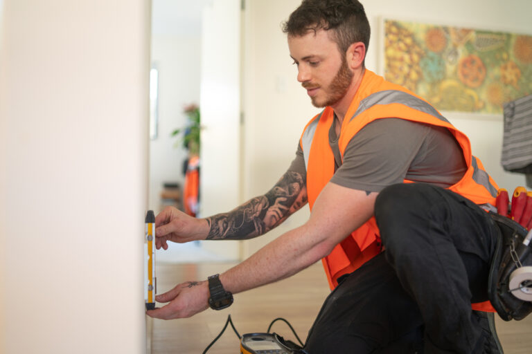 This image features a tradesperson wearing a bright orange safety vest and working indoors. They are using a measuring tool on a wall, with a focus on precision and care. The worker’s tool belt, visible on their hip, suggests expertise in installation or maintenance tasks, aligning with energy upgrades like air conditioning or heat pump installations under programs like the Victorian Energy Upgrade. The background displays a cozy, home-like setting, reinforcing the residential focus of these energy efficiency rebates and improvements.