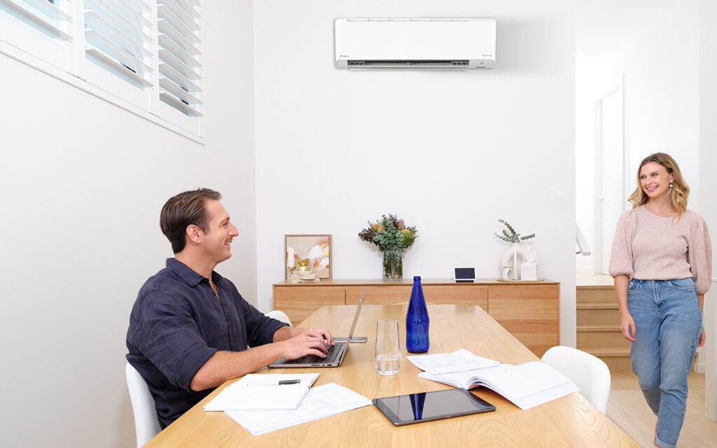 Modern home with a Daikin split system air conditioner installed on the wall, providing energy-efficient heating and cooling. A man working on his laptop and a woman entering the room, enjoying a comfortable indoor climate. Ideal air conditioning solution for Melbourne homes.